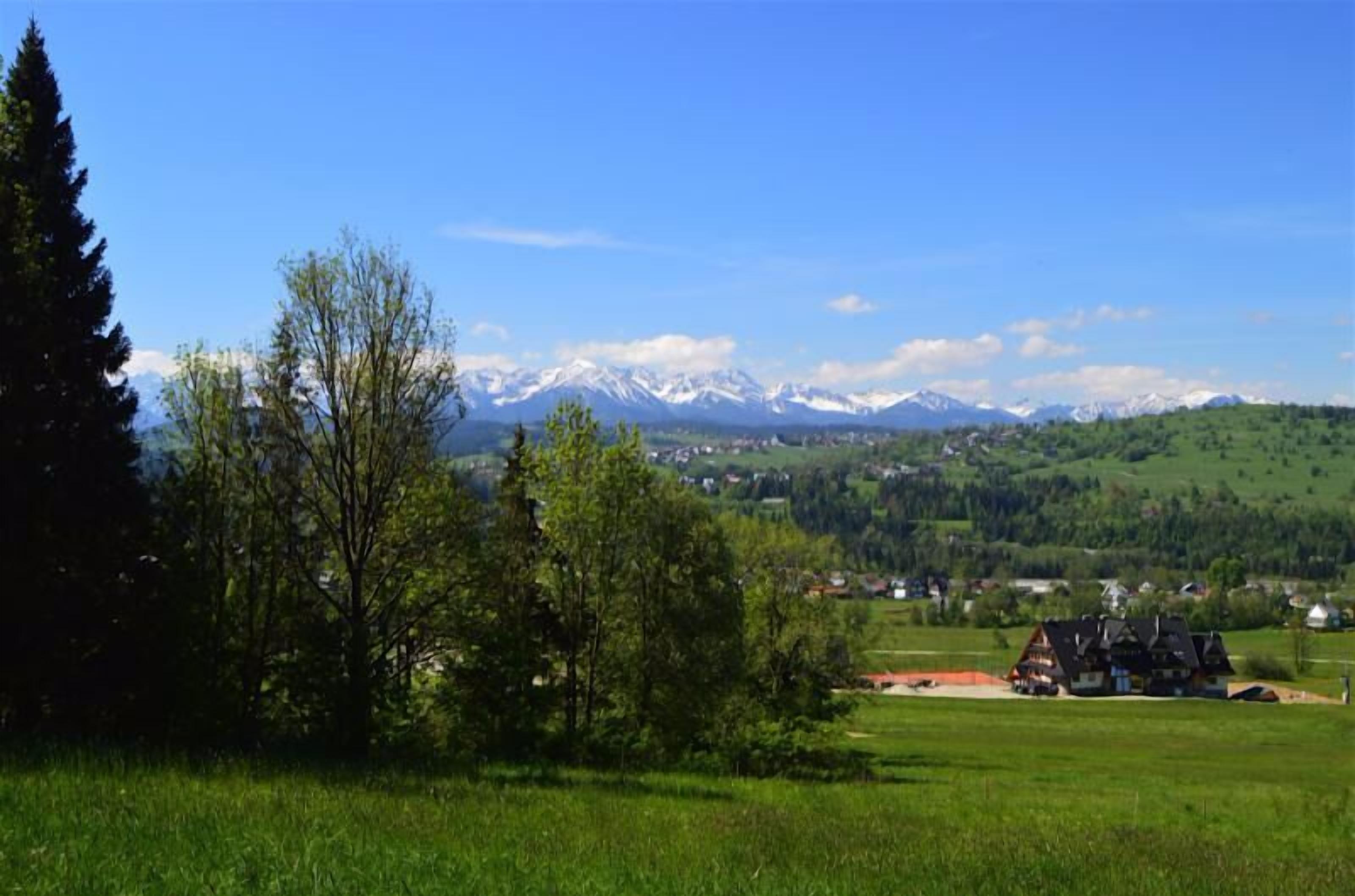 A rural landscape with a house, green fields, and snow-capped mountains in the background.