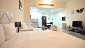 A hotel room with a large bed, a desk, a chair, a television, and a kitchenette in the background.