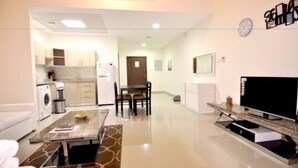 A modern living room with a flat-screen TV, a dining area with a table and chairs, and a kitchen with a washing machine and a refrigerator.