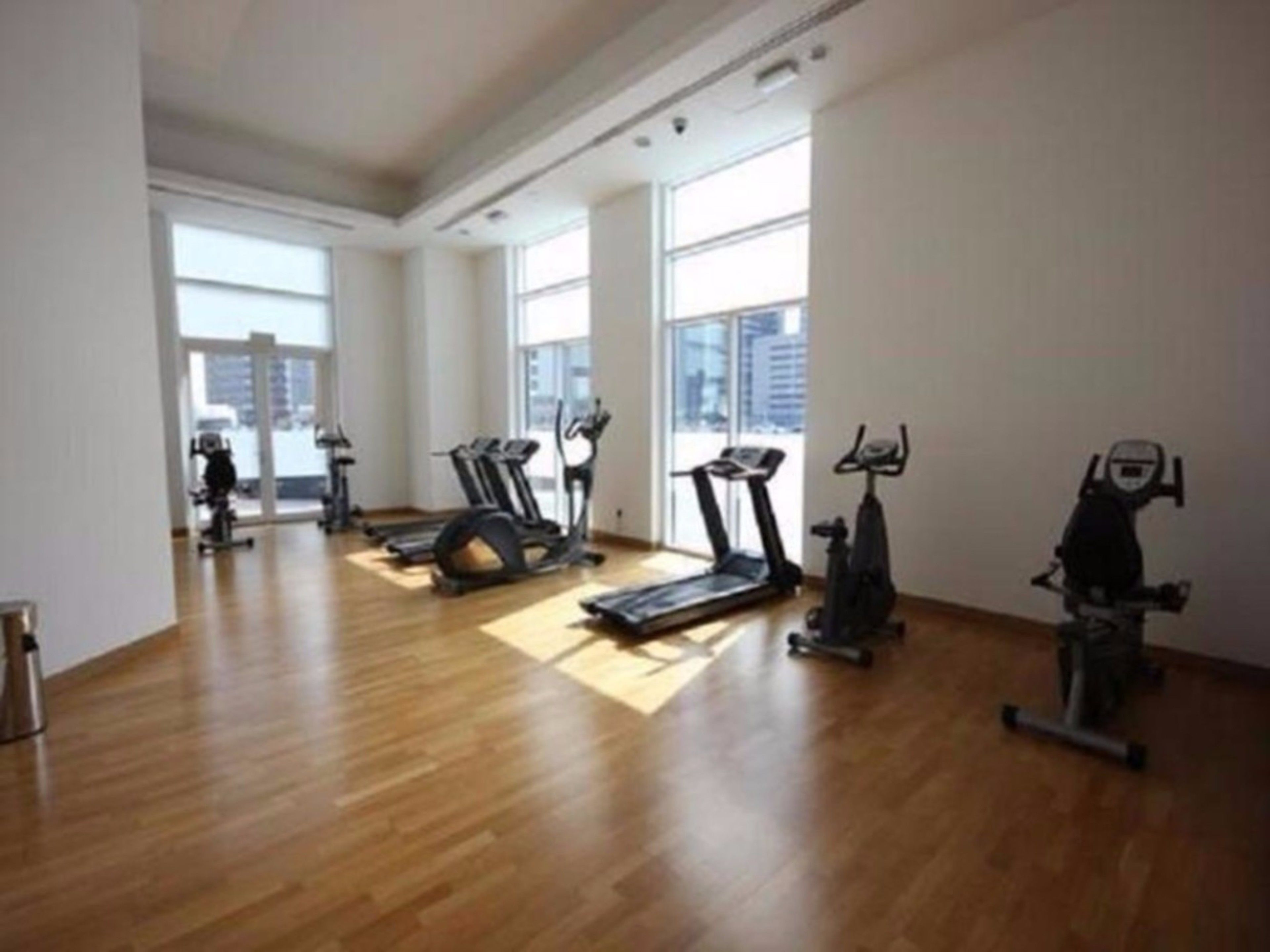 A spacious gym with multiple exercise machines, large windows, and a wooden floor.