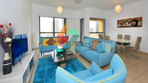 A modern living room with a blue sofa, a coffee table, and a TV. There is a dining area with a table and chairs. - Piks Key - Sadaf 4 (Dubai)