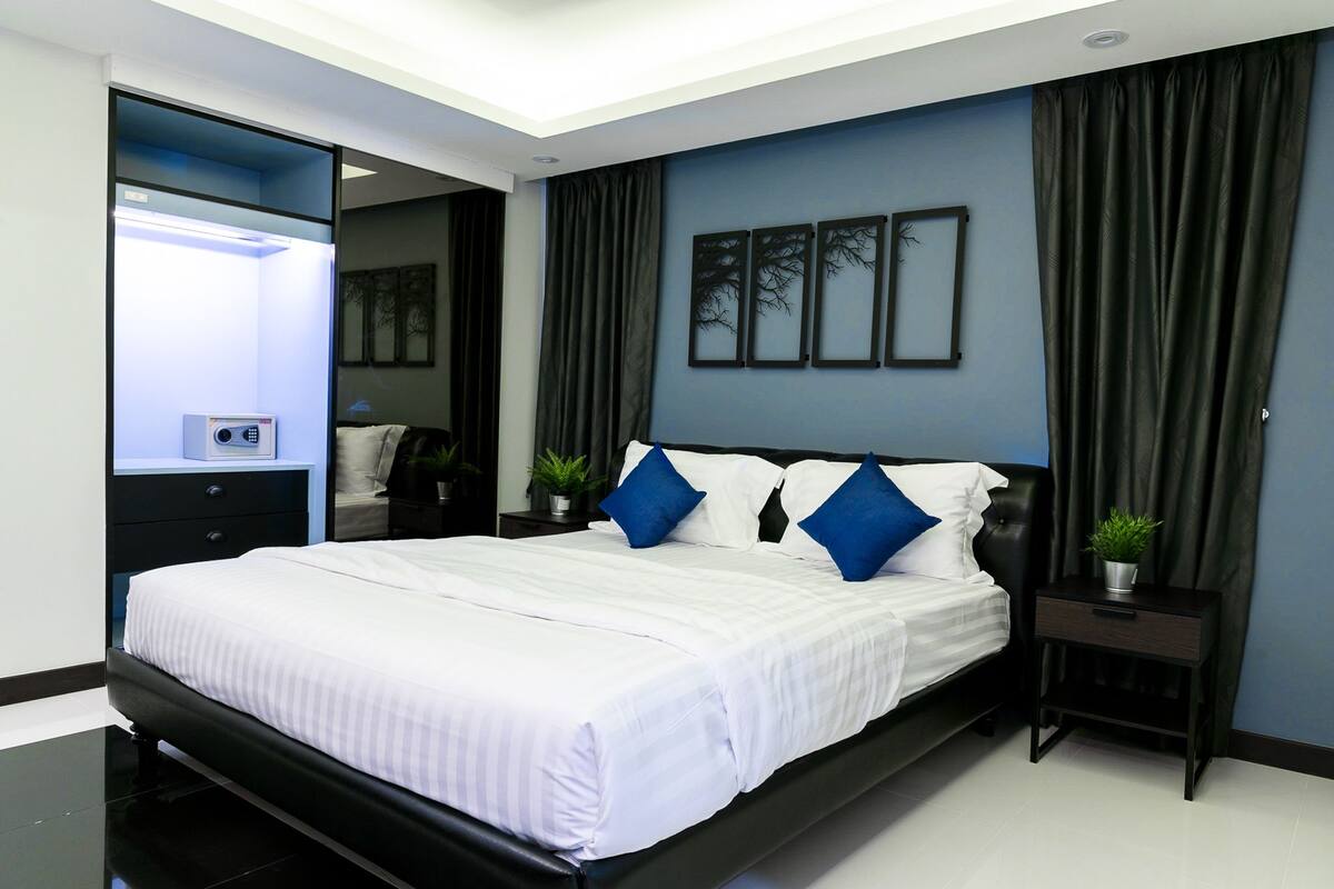 A modern bedroom with a black bed frame, white bedding, and blue accent pillows. There is a small bedside table with a plant and a nightstand with a lamp.