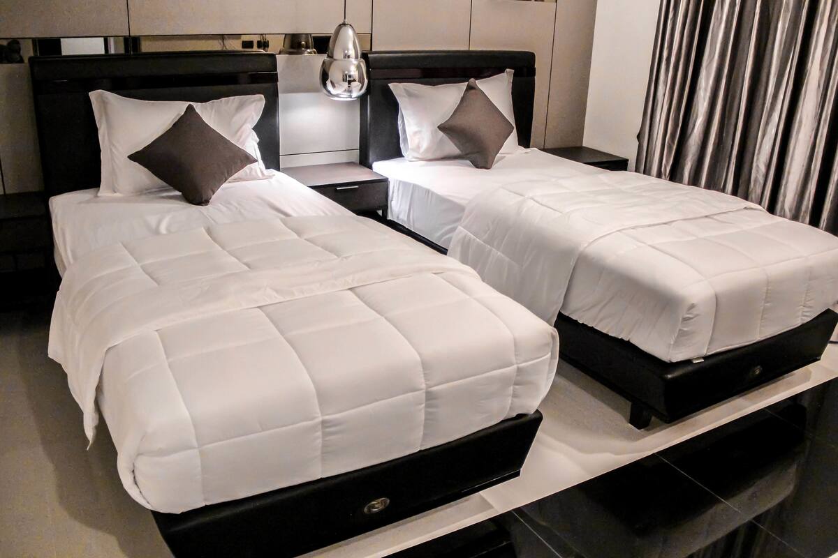 A hotel room with two beds, each with white bedding and dark headboards. There is a nightstand with a lamp between the beds.