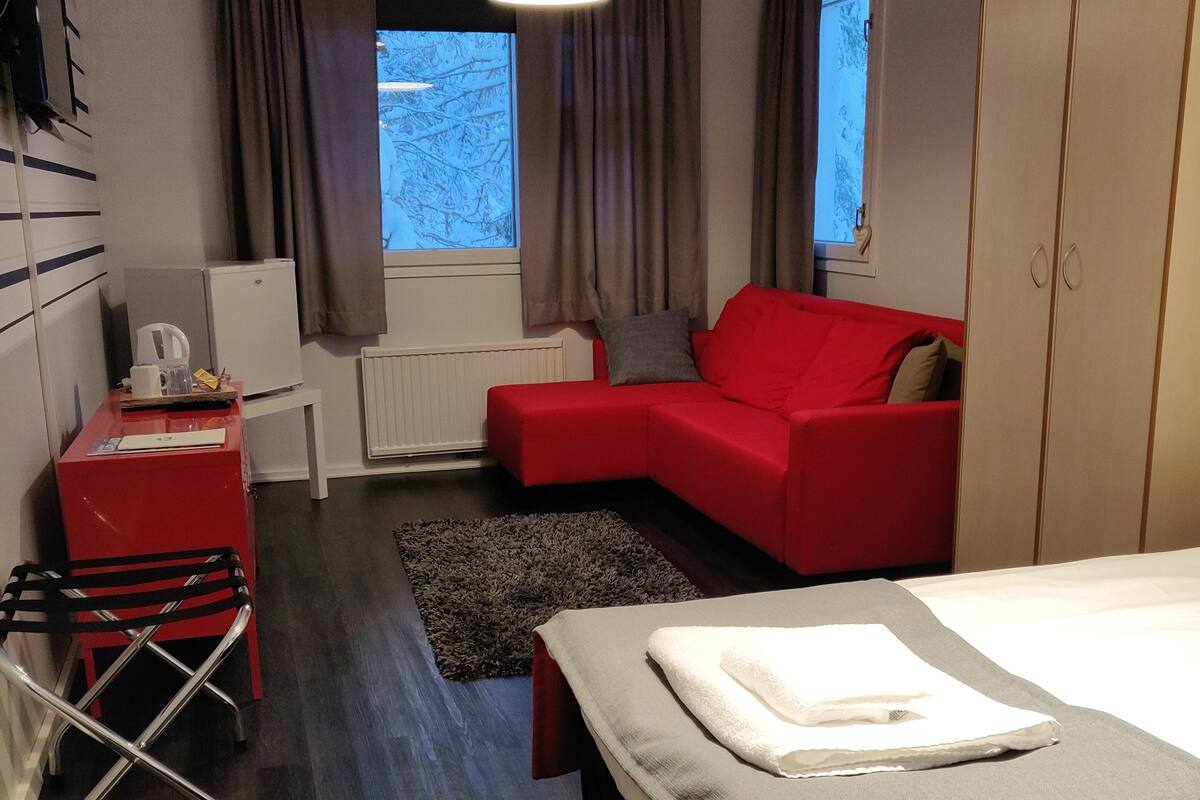 Une chambre d’hôtel moderne avec un canapé rouge, un petit bureau avec une bouilloire, un lit recouvert d’une couverture blanche et une vue sur les arbres par la fenêtre.