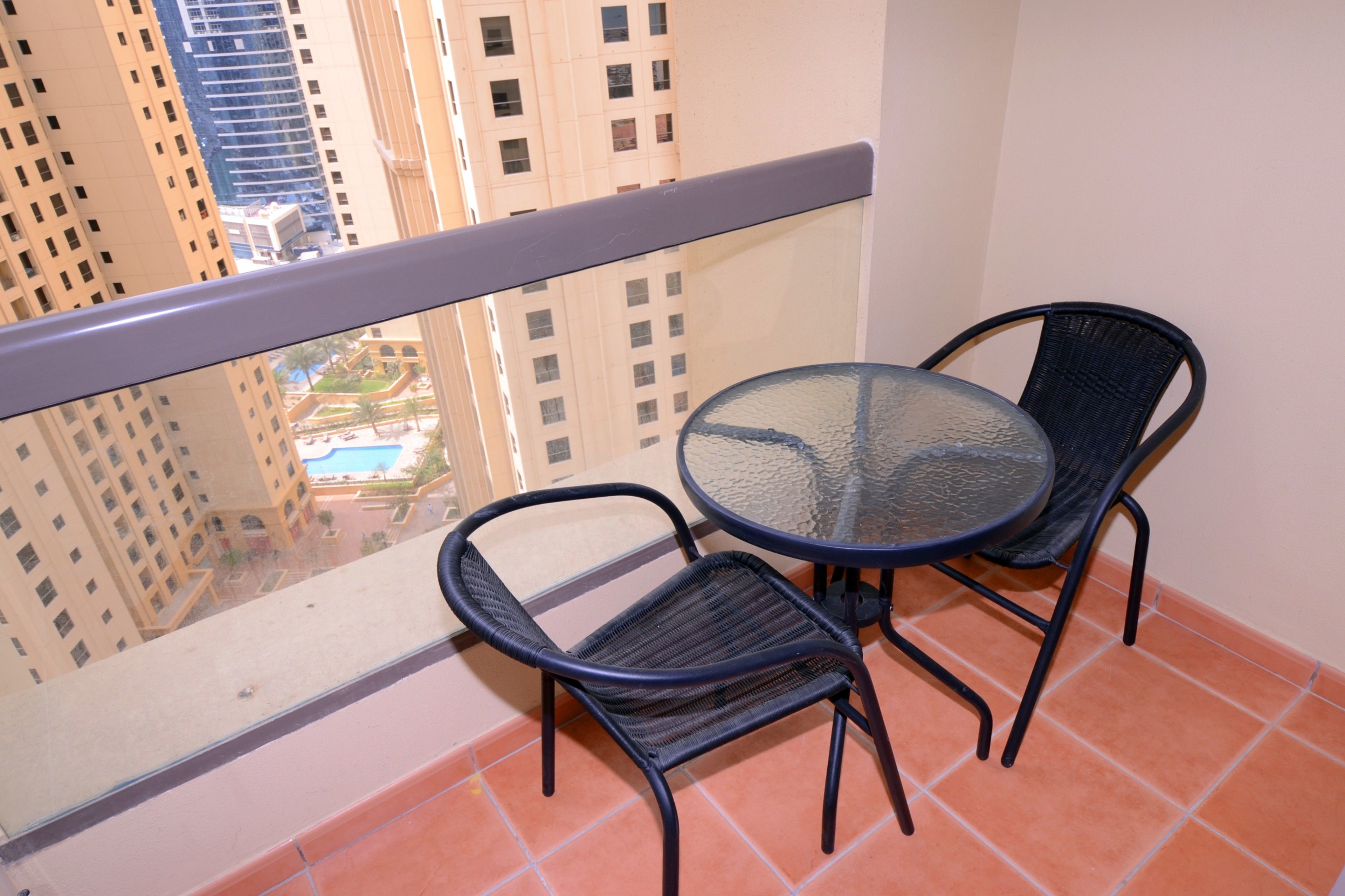 Un balcón con una mesa de vidrio y dos sillas, con vistas a un paisaje urbano con edificios altos.