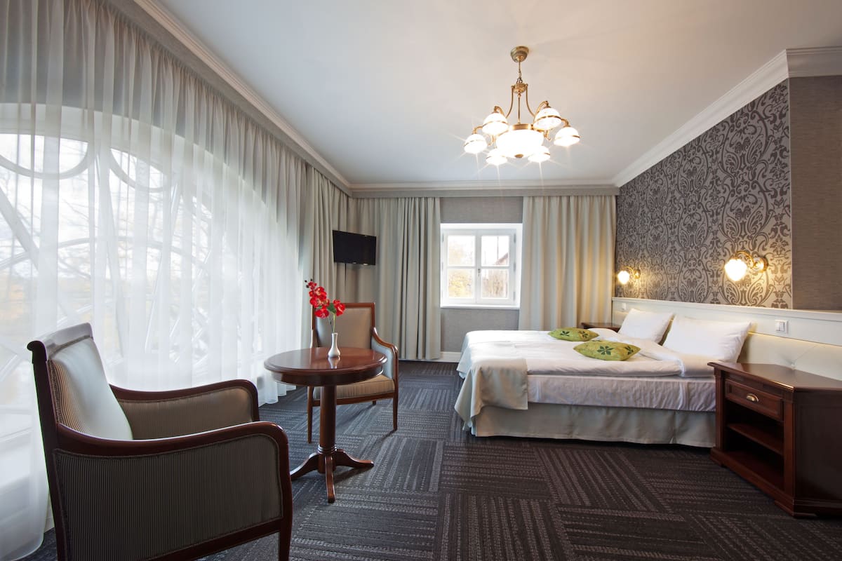 A hotel room with a bed, a chair, a table, a TV, and a chandelier.