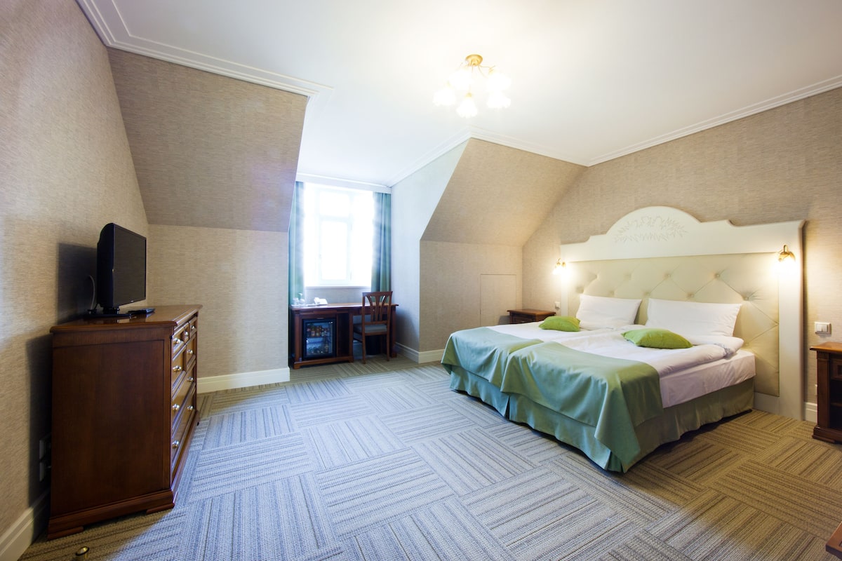 A bedroom with a large bed, a desk with a chair, a television on a dresser, and a window with curtains.