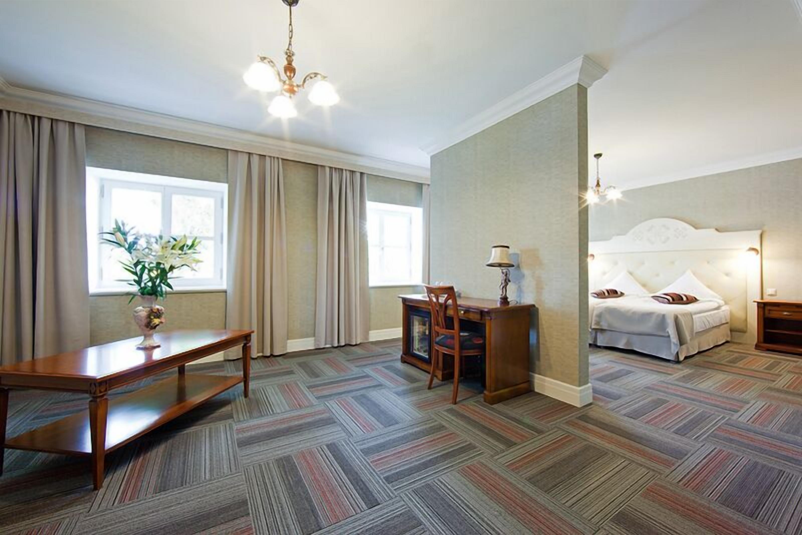 Habitación de hotel con una cama grande, un escritorio con una silla, una mesita con una lámpara y una planta en maceta junto a la ventana.