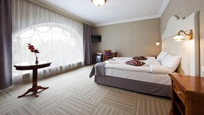 Ett hotellrum med en säng, ett litet bord med en vas, en stol, ett skrivbord och en tv.