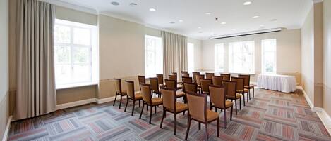 Een vergaderzaal met rijen stoelen, grote ramen en een kamerbreed tapijt met patroon.