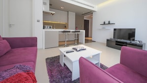A modern living room with a white coffee table, purple sofas, a flat-screen TV, and a kitchen area with a bar. - Piks Key - West Avenue (Dubai)
