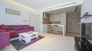 A modern living room with a purple sofa, white coffee table, and a kitchen area with a microwave and bar stools. - Piks Key - West Avenue (Dubai)
