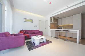 A modern living room with a purple sofa, a white coffee table, and a kitchen area with a microwave and bar stools.