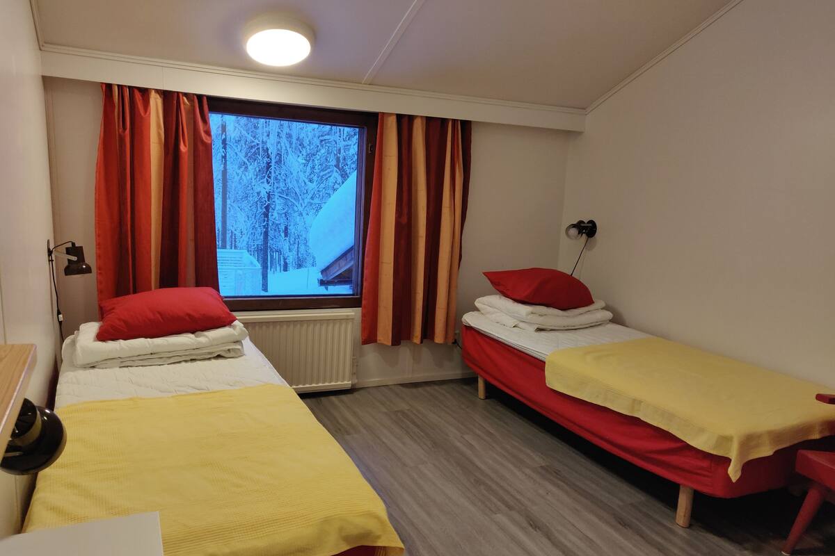 A hotel room with two single beds, a window with curtains, a bedside table, and a desk with a lamp.