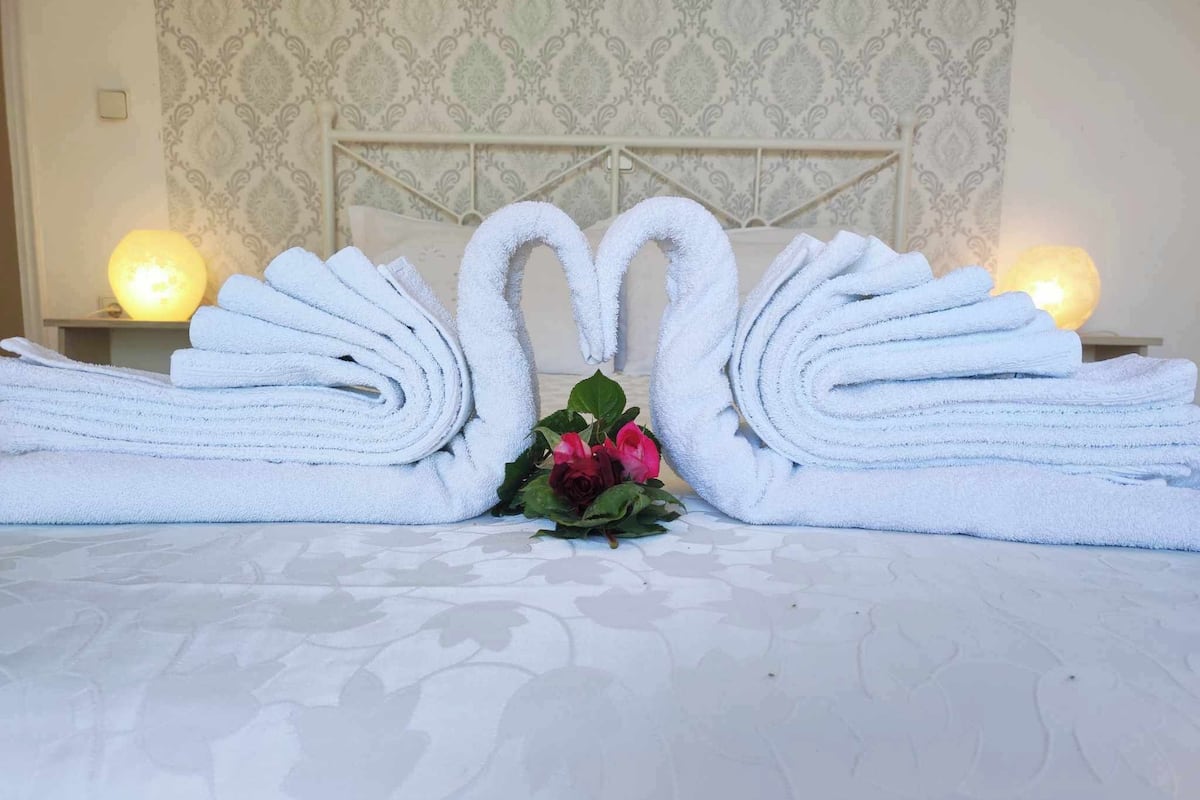 A neatly made bed with towels arranged in the shape of swans, a bouquet of flowers, and a patterned wallpaper background.