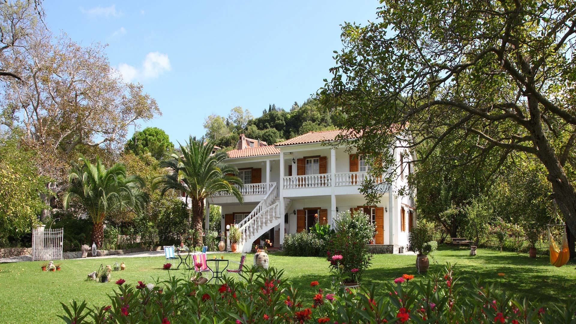 A two-story white building with a red-tiled roof, surrounded by lush greenery and a well-maintained garden.