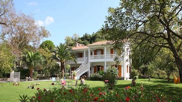A two-story white building with a red-tiled roof, surrounded by lush greenery and a well-maintained garden.