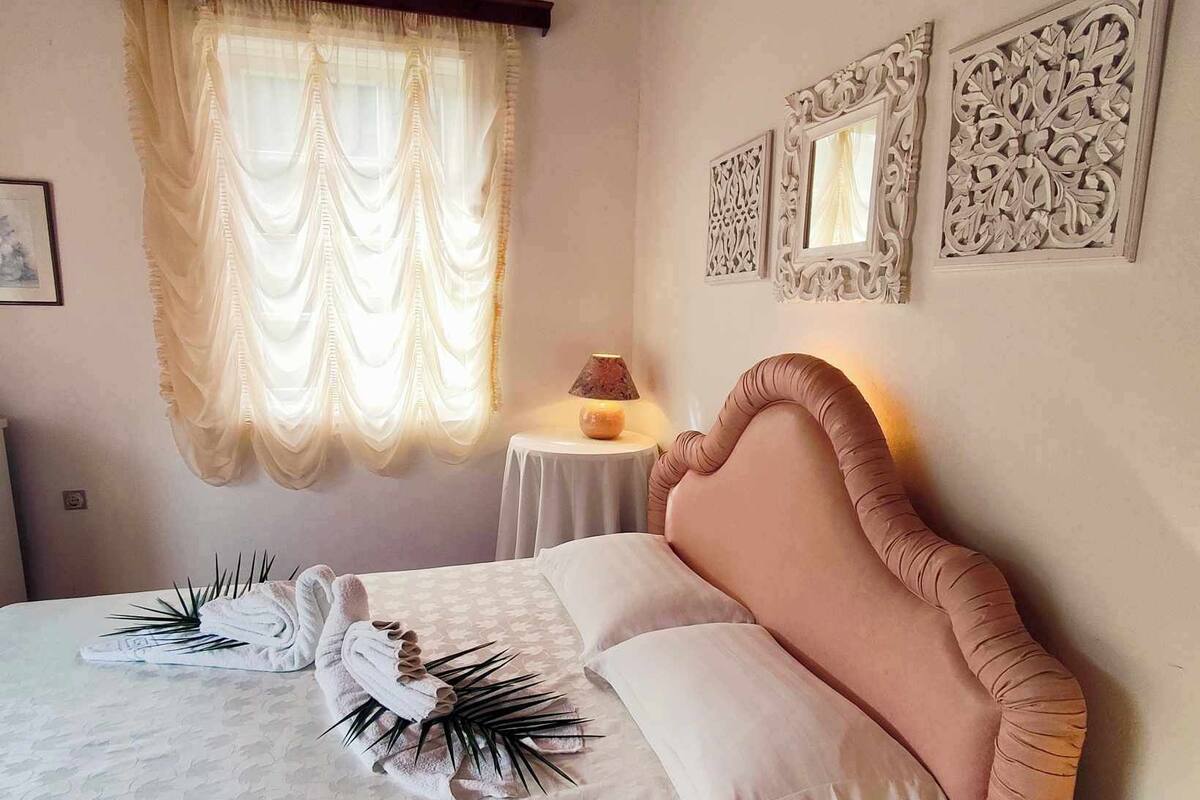 A neatly made bed with a pink headboard, white linens, and decorative pillows. There is a small round table with a lamp beside the bed.