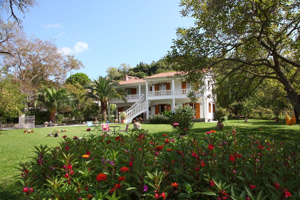A two-story building with a white exterior and red roof, surrounded by lush greenery and flowering plants.