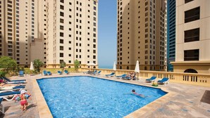 A swimming pool surrounded by lounge chairs and umbrellas, with tall residential buildings in the background. - Piks Key - Bahar 1 (Dubai)