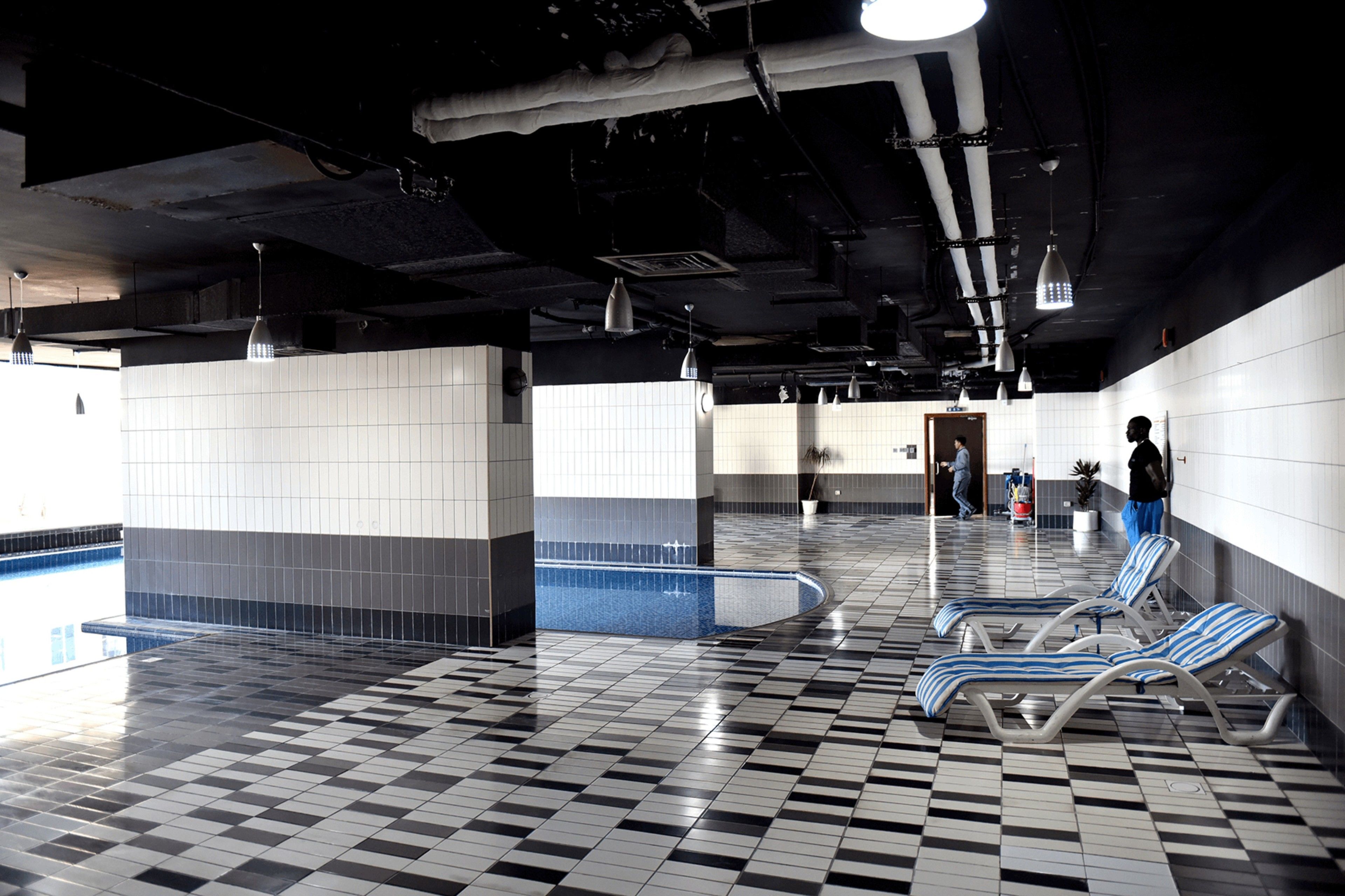 A pool area with tiled flooring, lounge chairs, and a tiled wall.