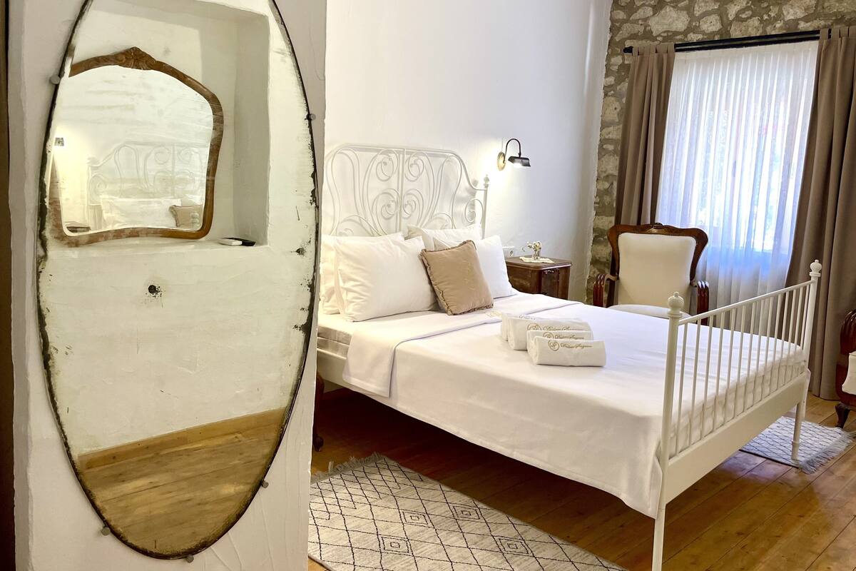 A bedroom with a white bed, a wooden floor, a stone wall, and a window with curtains.