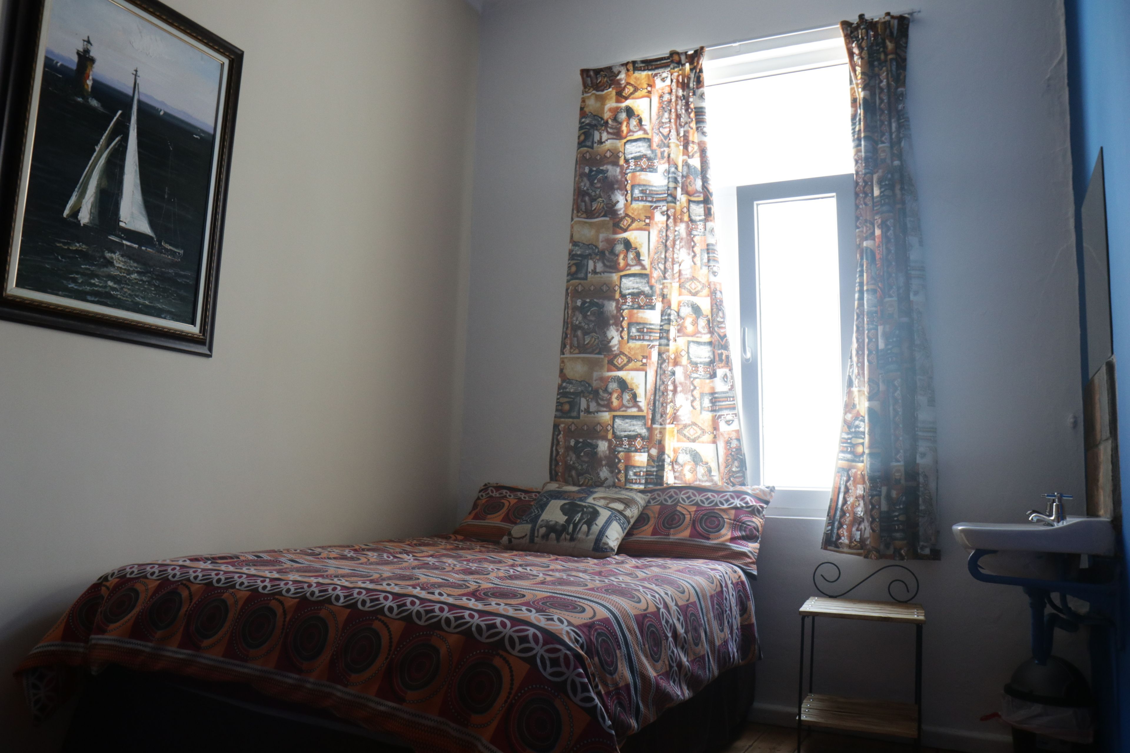 A bedroom with a bed, a window with patterned curtains, a small bedside table, and a wall-mounted sink.