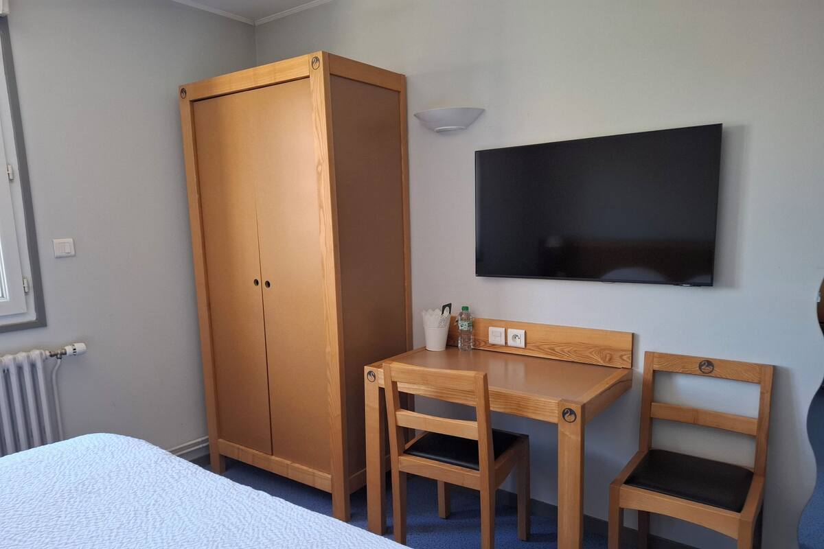 A bedroom with a bed, a wooden wardrobe, a desk with two chairs, a television, and a radiator.