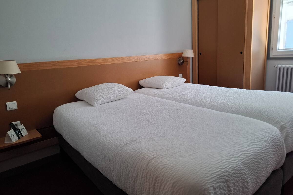 A hotel room with two beds, wooden headboard, bedside lamps, a nightstand with a phone and papers, and a window with blinds.