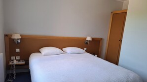 Ein ordentlich gemachtes Bett mit zwei Kissen, einem Holzkopfende, Nachttischlampen, einem Nachttisch mit Telefon und Buch, und einer Tür rechts.