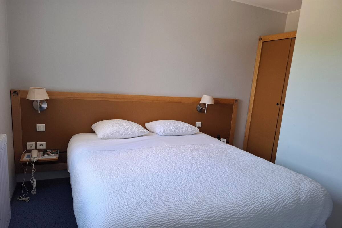 A neatly made bed with two pillows, a wooden headboard, bedside lamps, a nightstand with a phone and a book, and a door to the right.