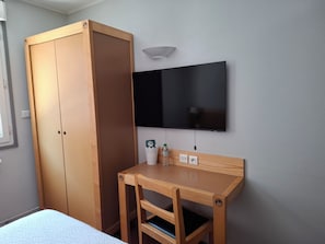 Ein Schlafzimmer mit einem Bett, einem Holzschrank, einem kleinen Holztisch mit Stuhl, einem flachbildfernseher an der Wand und einer Wandleuchte.