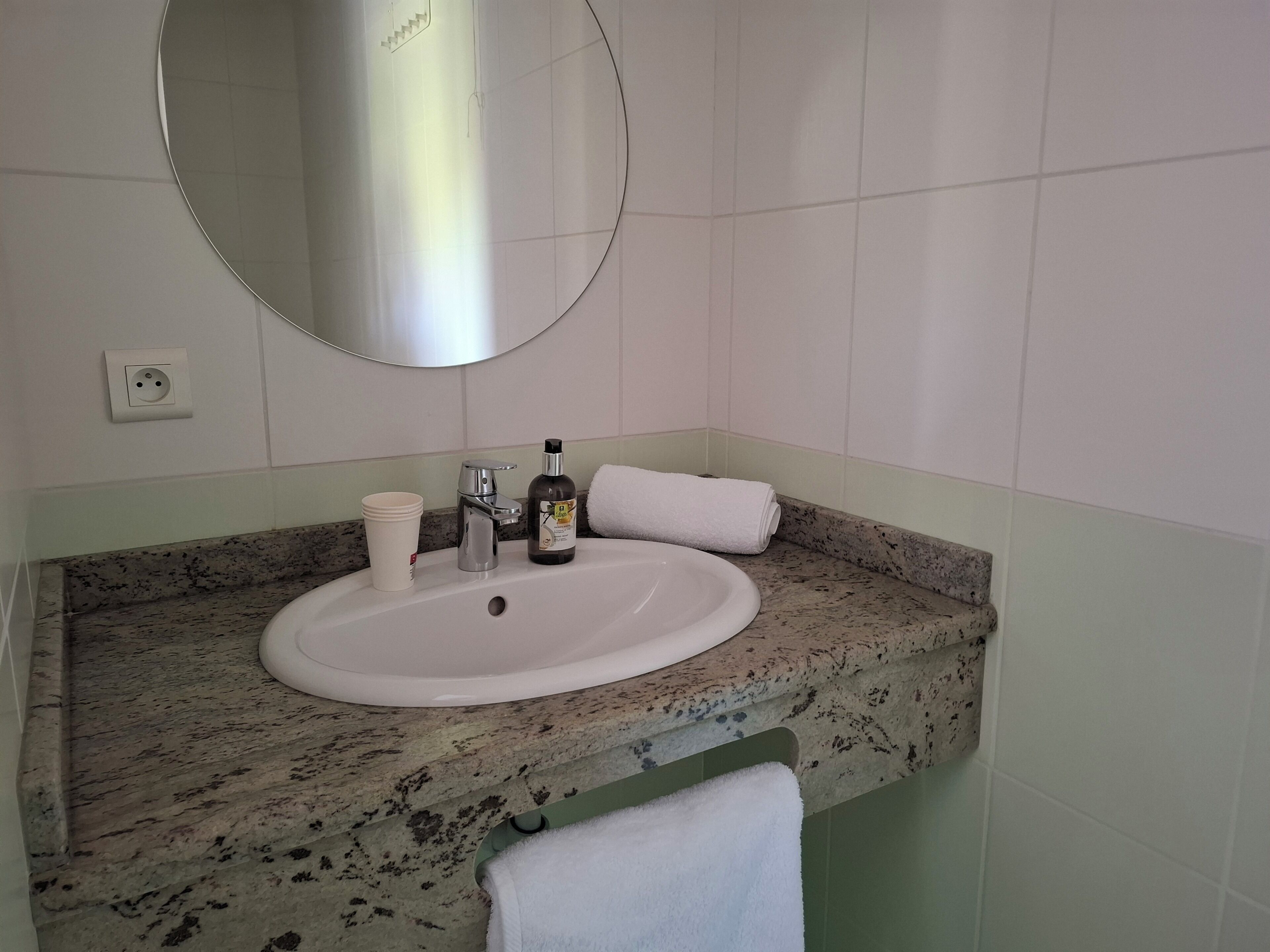 Une salle de bain avec un lavabo blanc, un miroir, un robinet, une bouteille et une tasse.