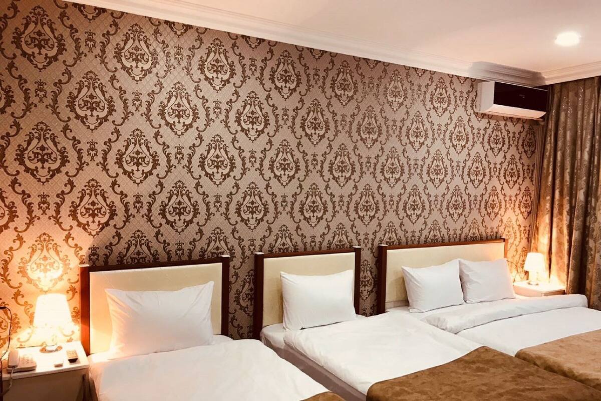Una habitación de hotel con tres camas, un papel tapiz estampado, un equipo de aire acondicionado y una lámpara de noche.