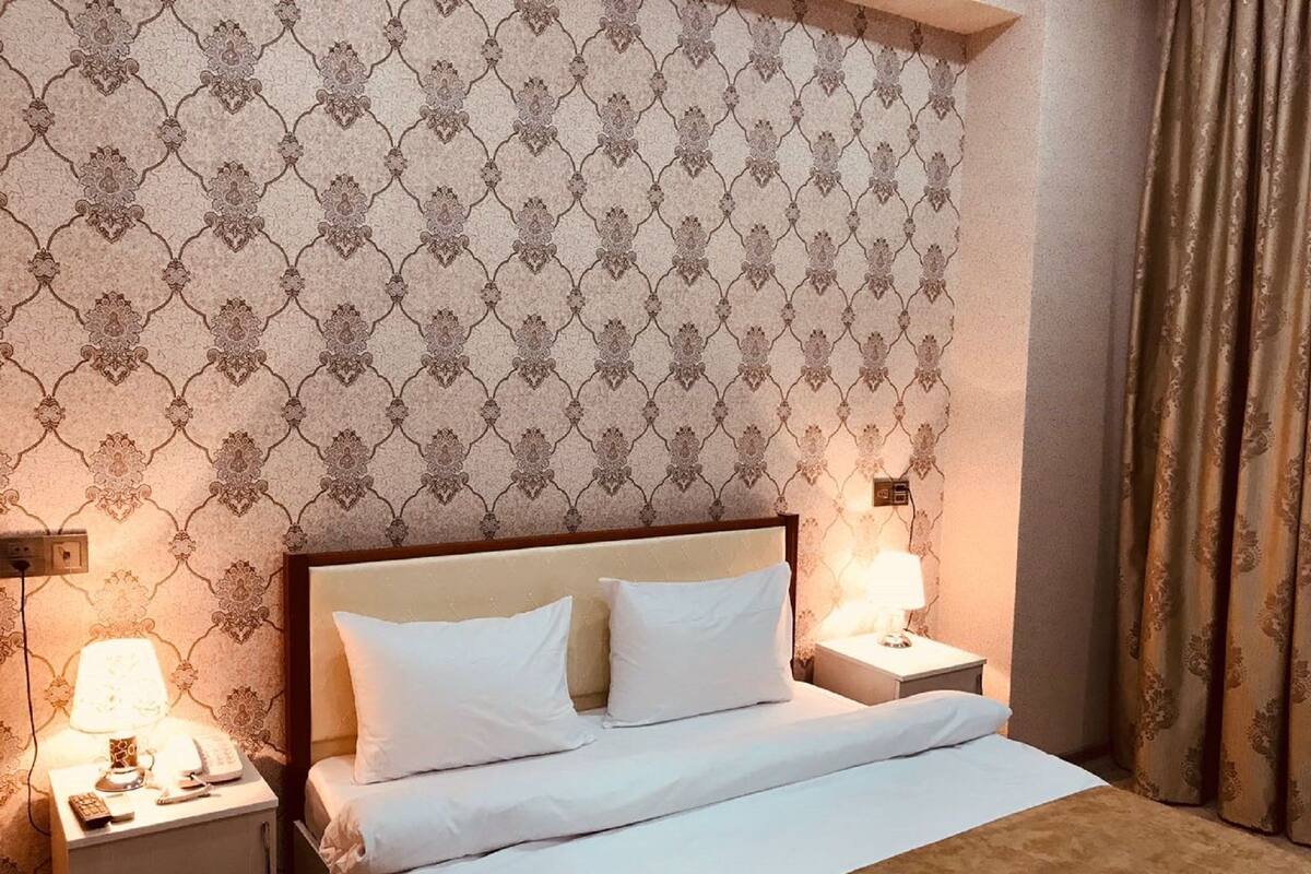 Una cama bien tendida con sábanas blancas, dos lámparas de noche y un papel tapiz estampado.