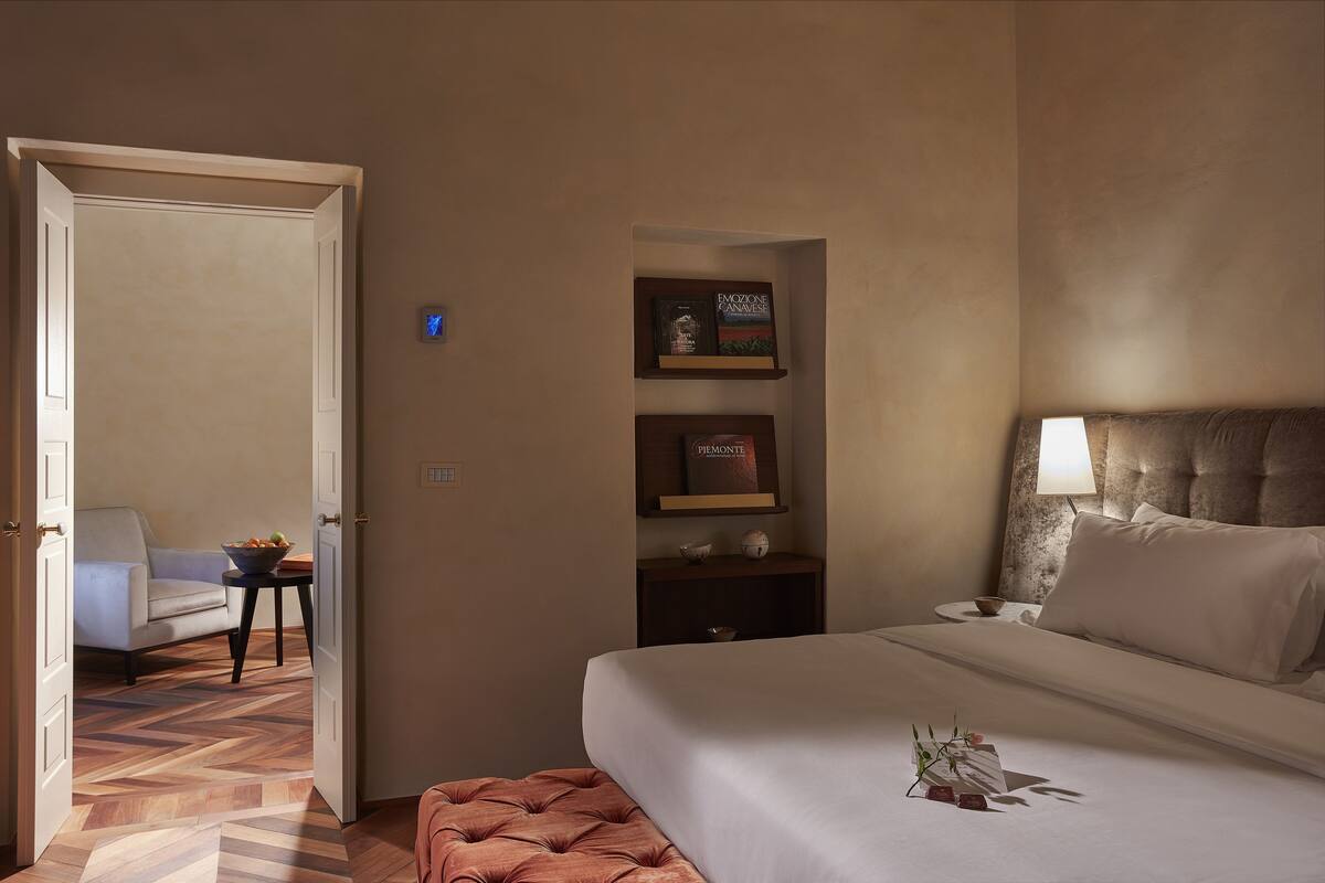 A hotel room with a bed, a chair, a small table with a bowl of fruit, and a shelf with decorative items.