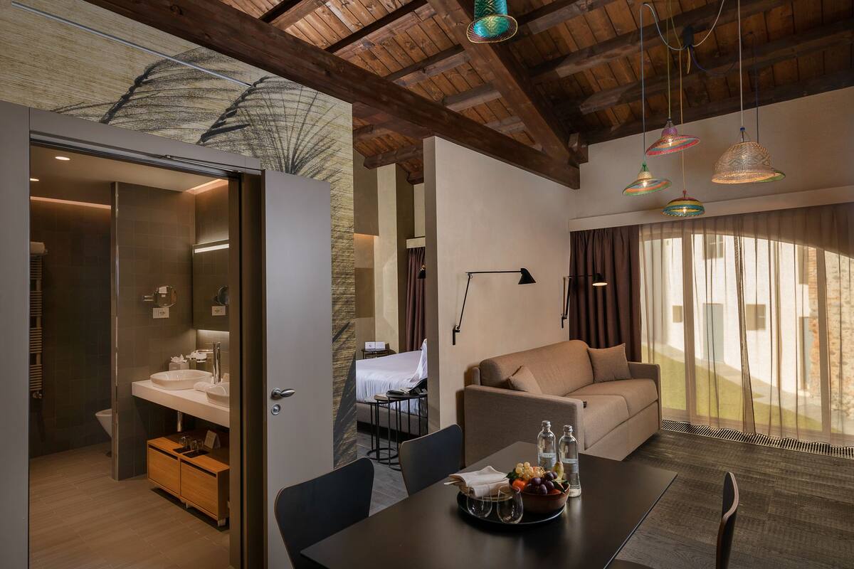 A modern hotel room with a wooden ceiling, a dining area with a table set for two, and a cozy seating area with a sofa.