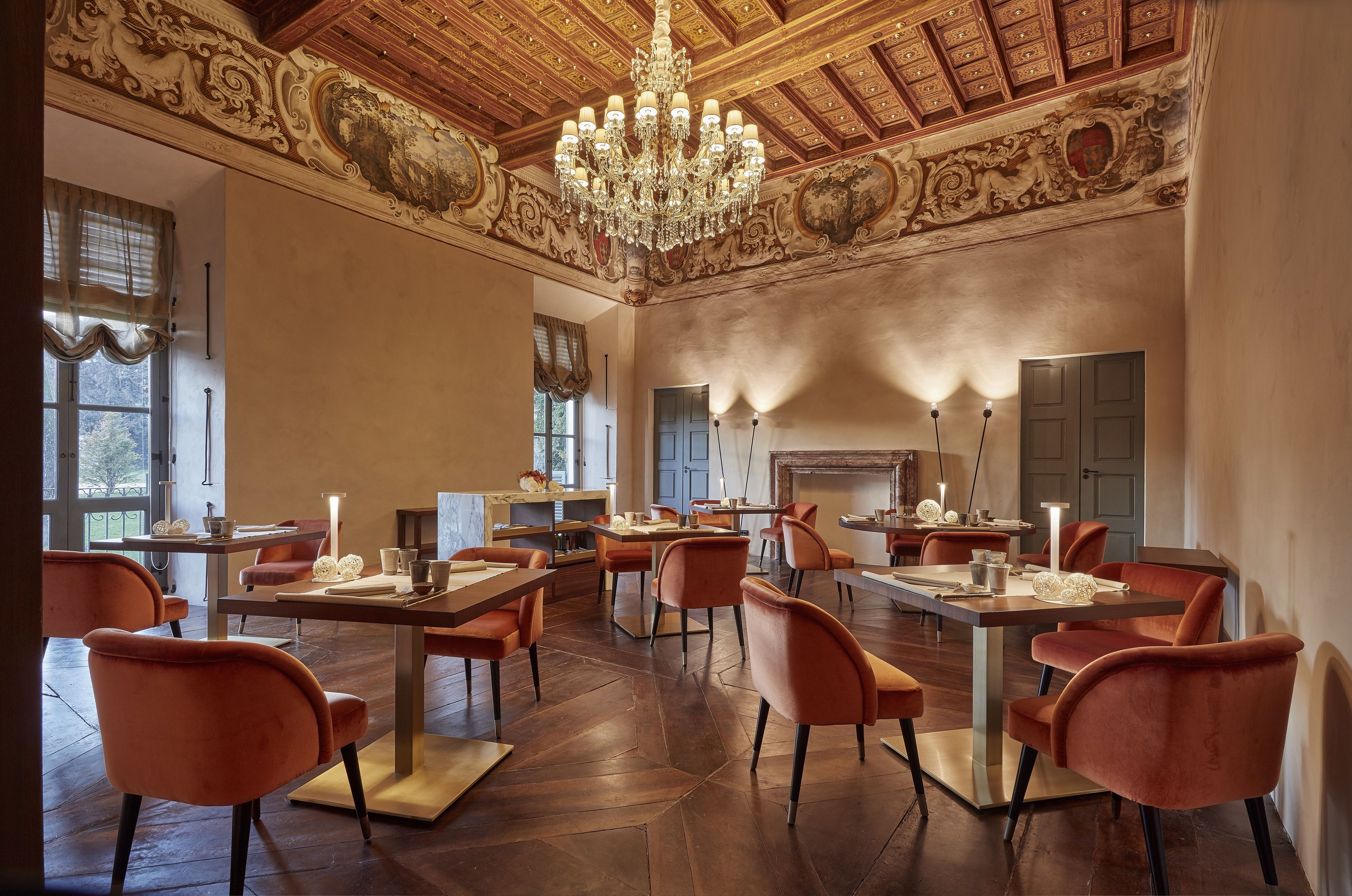 A dining area with ornate ceiling and chandelier, wooden flooring, and orange upholstered chairs.