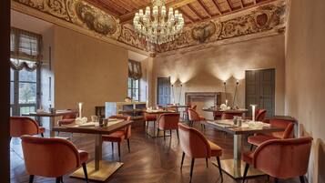 A dining area with ornate ceiling and chandelier, wooden flooring, and orange upholstered chairs.