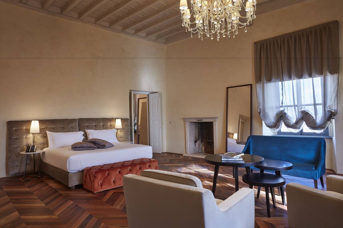 A spacious hotel room with a large bed, a fireplace, a chandelier, and a view of the outdoors.