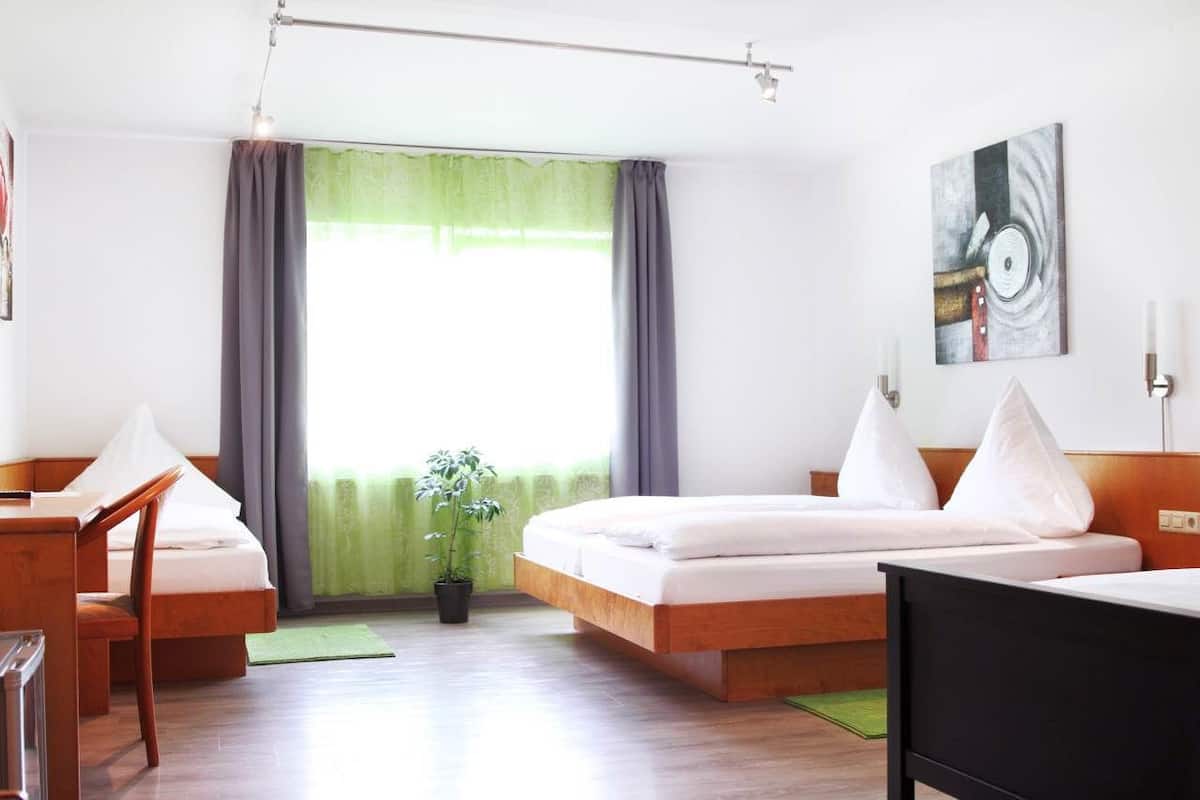 Habitación de hotel moderna con una cama de madera, un escritorio y una silla. Hay una ventana con cortinas, una planta y un cuadro en la pared.