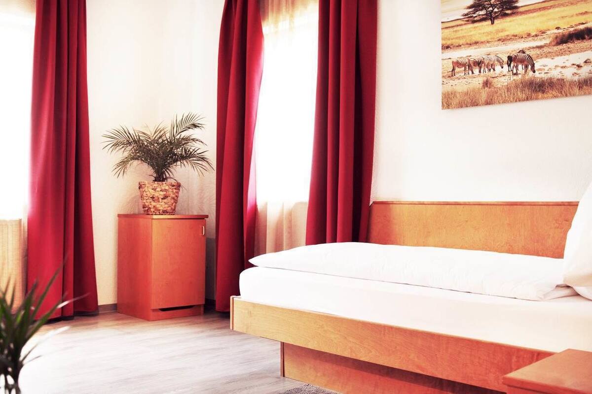 Un dormitorio con cama de madera, cortinas rojas, una mesita de noche con una planta y un cuadro de cebras en la pared.