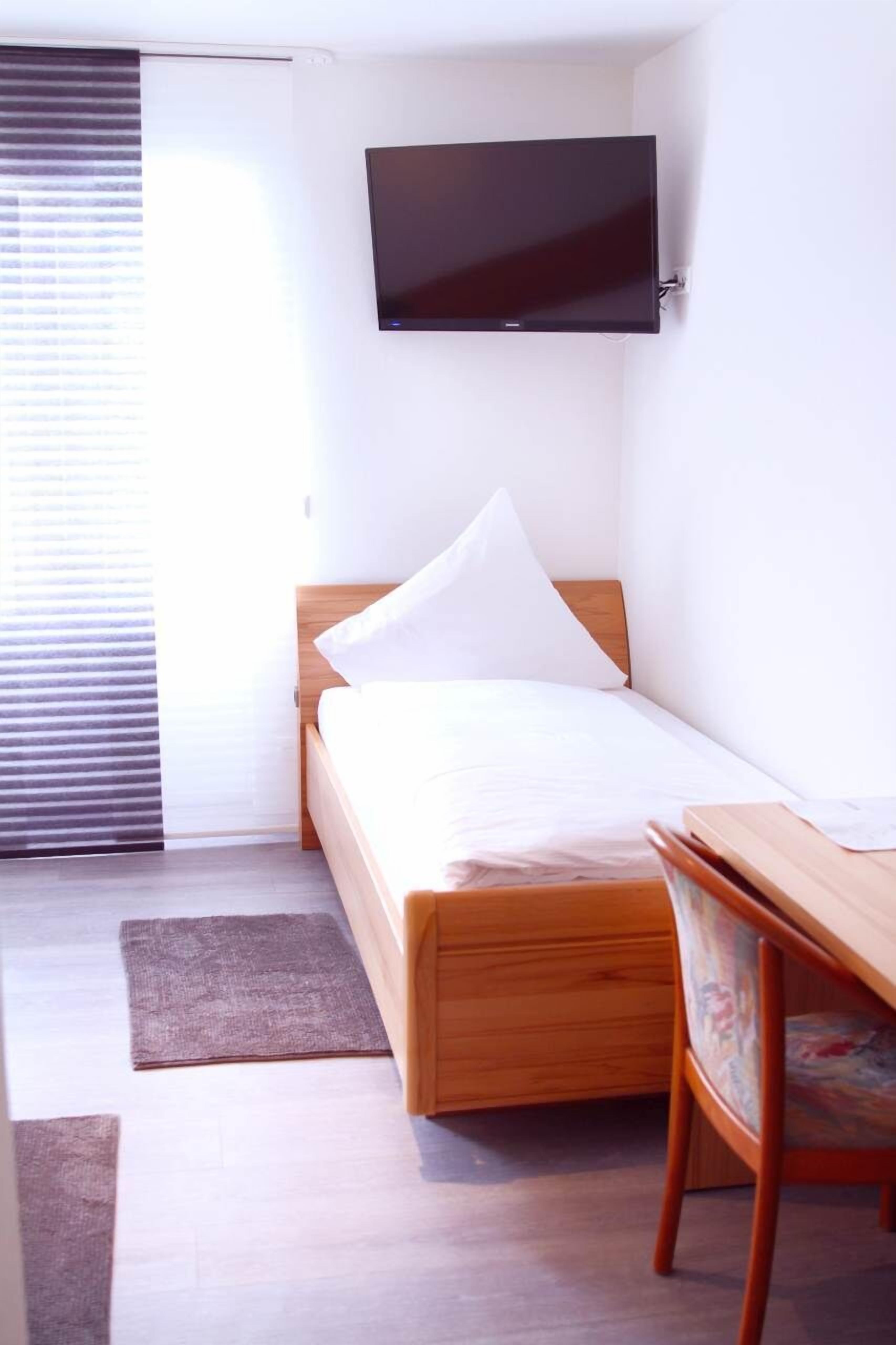 A small, neatly arranged room with a bed, a desk, and a mounted TV.