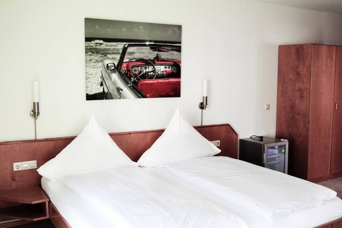 Habitación de hotel con cama, almohadillas blancas, cabecera de madera, un cuadro colgado en la pared que muestra un coche, armario de madera y un pequeño calentador.