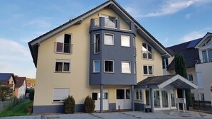 Ein mehrgeschossiges Wohngebäude mit einem Balkon im zweiten Stock, einem überdachten Eingang und einer kleinen Gartenanlage.