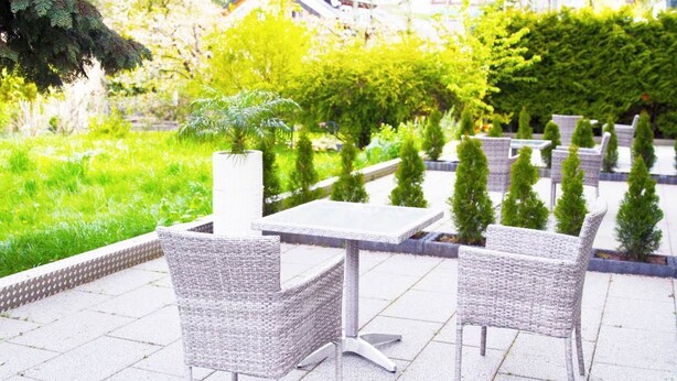 Eine Terrasse im Freien mit Korbmöbeln und einem weißen Tisch, umgeben von Grünpflanzen und einer Hecke.