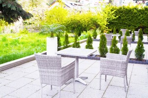 Un patio al aire libre con mobiliario de mimbre y una mesa blanca, rodeado de follaje y un seto.
