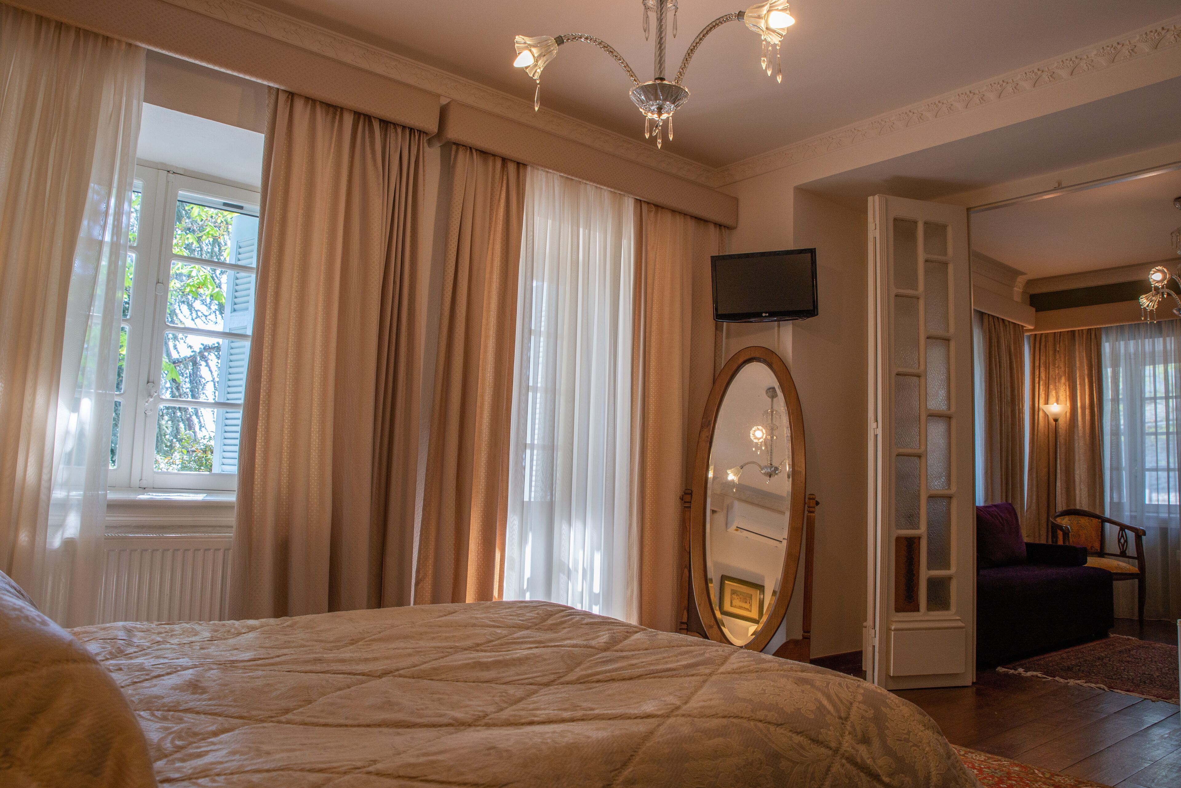 Una camera da letto con un letto grande, uno specchio, una televisione e vista sull'esterno attraverso una finestra con tende.