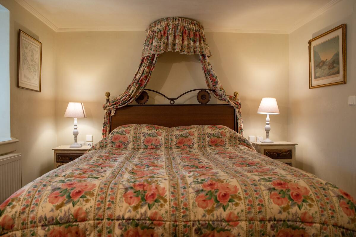 Un dormitorio con colcha estampada, cama con dosel, dos mesitas de noche con lámparas y cuadros enmarcados en las paredes.