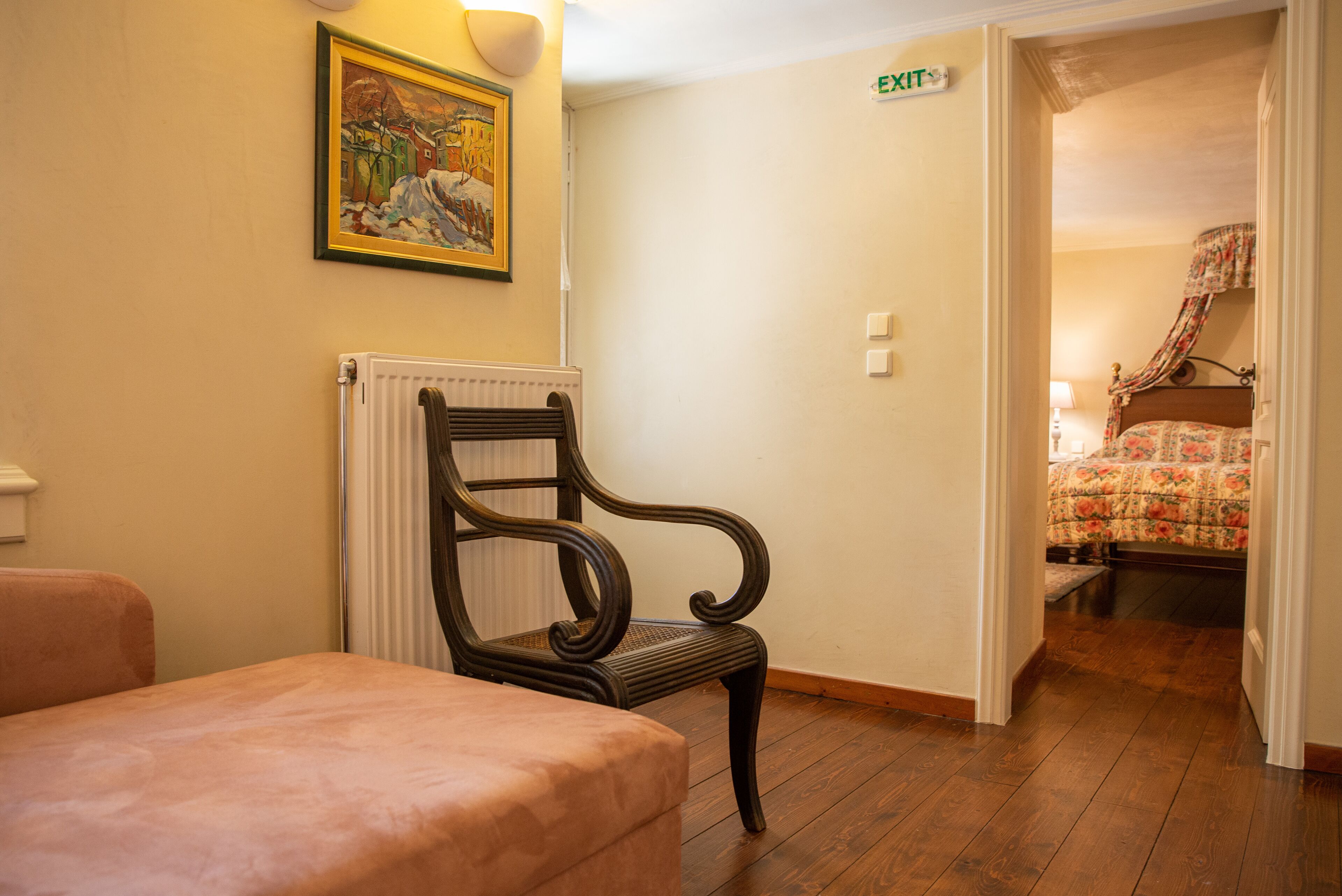 A hotel room with a wooden chair, a radiator, a painting on the wall, and a bed with a floral bedspread.